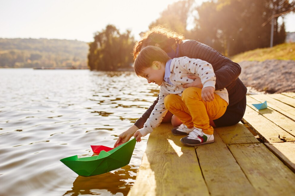 woman with little boy playing with toy paper ship near the lake